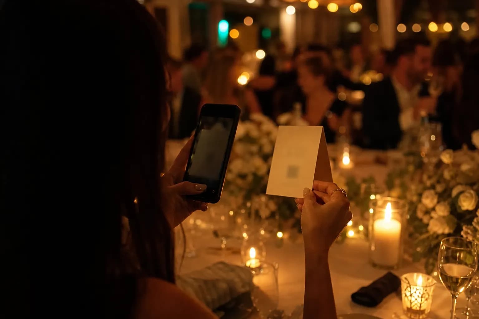 Ein Gast hebt auf einer warm beleuchteten Feier eine kleine Tischkarte an und richtet sein Smartphone darauf, im Hintergrund feiern Gäste. (KI-generiertes Bild)
